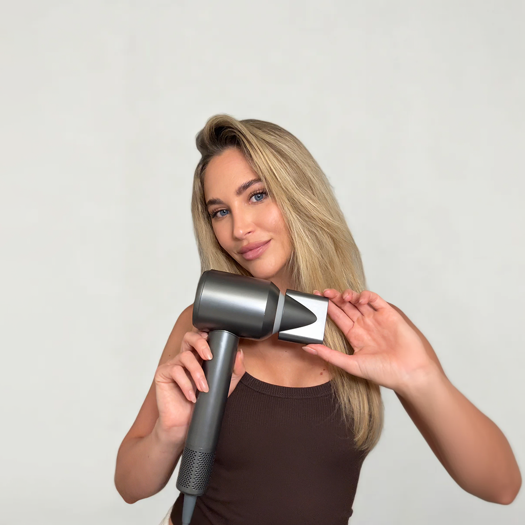 A woman with long blonde hair is holding a Vividreamore dark gray high-performance hair dryer with its nozzle attachment. She smiles softly at the camera, wearing a dark brown sleeveless top. The plain light-colored background highlights both the hair dryer and her hair’s smooth texture.